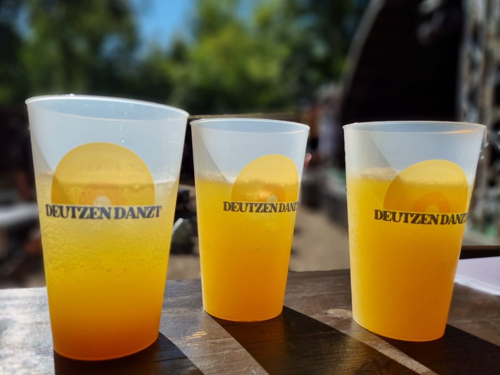 Drei DEUTZEN-DANZT-Becher mit gelben Getränken in der Sonne
